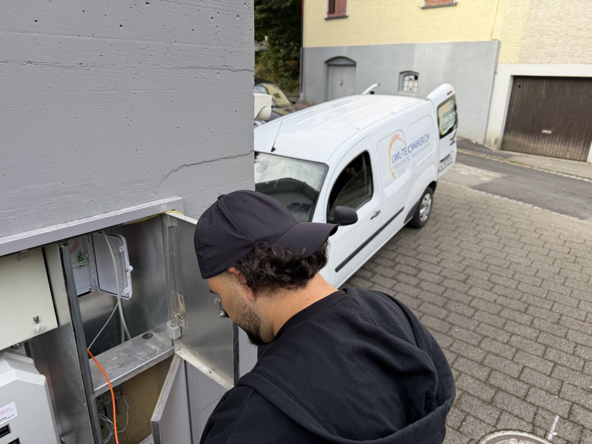Techniker am geöffneten Glasfaserverteiler an einer Hauswand, daneben das Montagefahrzeug