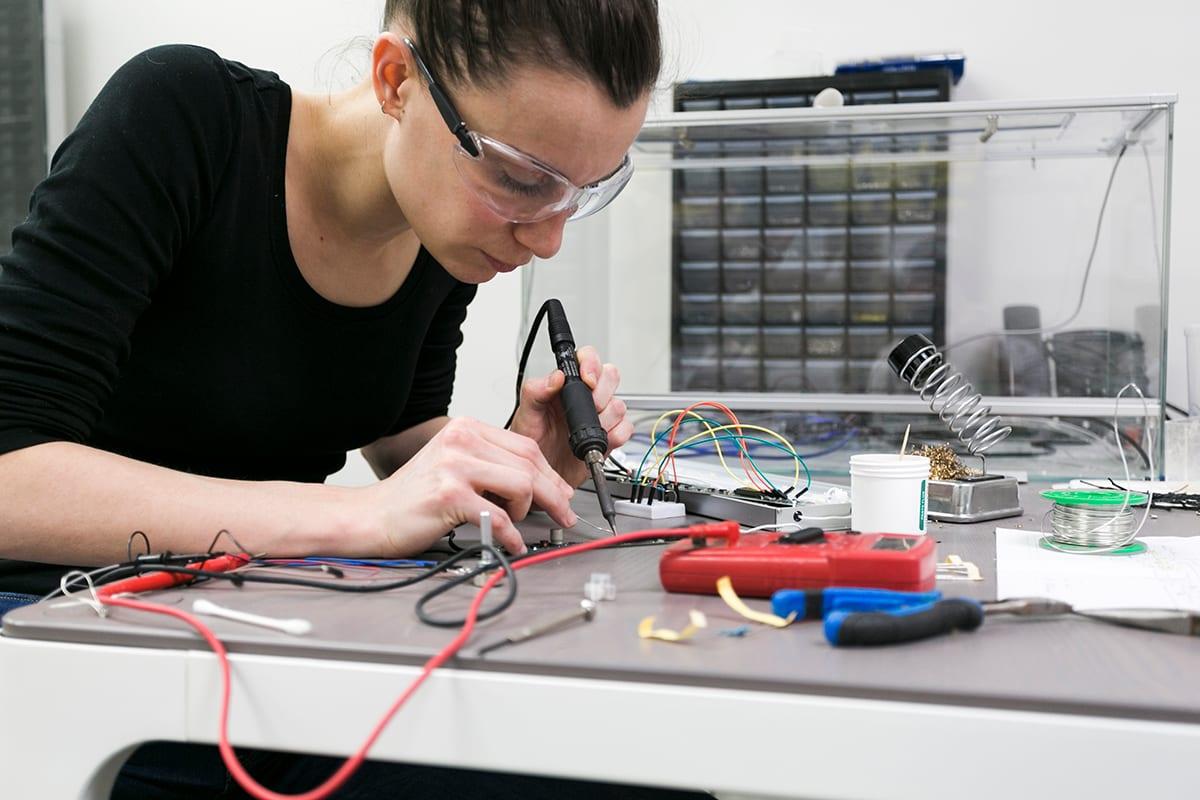 Mitarbeiterin lötet ein Bauteil am Arbeitsplatz in der Werkstatt